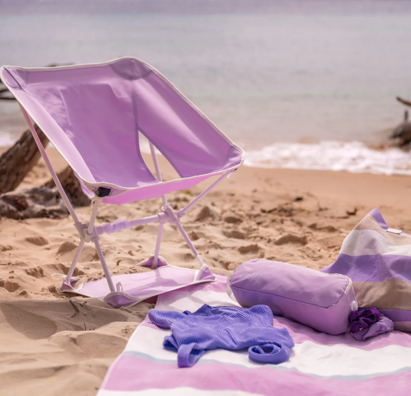 A lilac-colored Subulo beach chair with a matching pink sand mat, set up on the beach with a clear blue sky and white sand background. A grey backpack and a cushion are also visible in the foreground.
