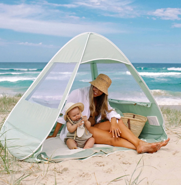 A sage green pop-up beach shelter with mesh windows set up on the beach, with an adult and a child inside.