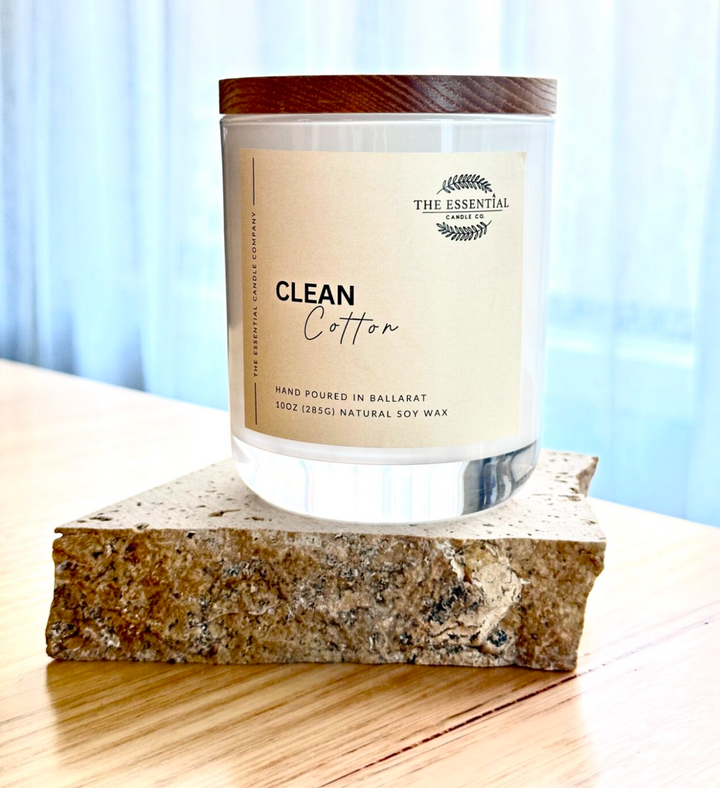 A clear soy candle with a beige label that reads 'Clean Cotton' and is branded by The Essential Candle Co. The candle is placed on a rocky surface with a blurred background.