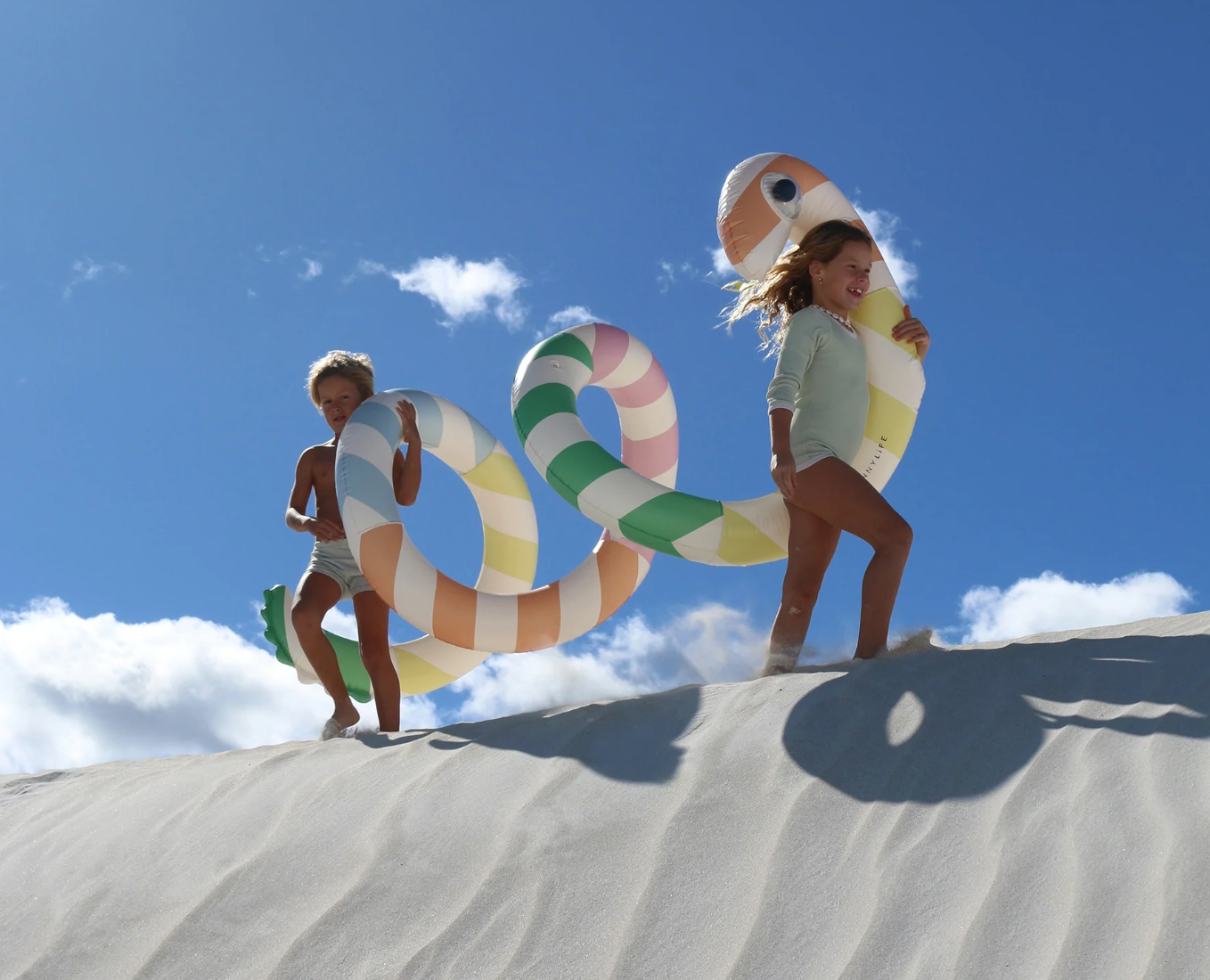 Two children playing with a large inflatable snake on a sandy dune under a blue sky.